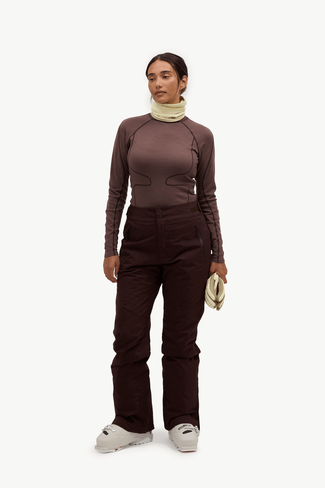 A model stands against a plain white studio background wearing a dark brown long-sleeve technical top, matching dark brown pants, a light beige neck gaiter, and white sneakers.