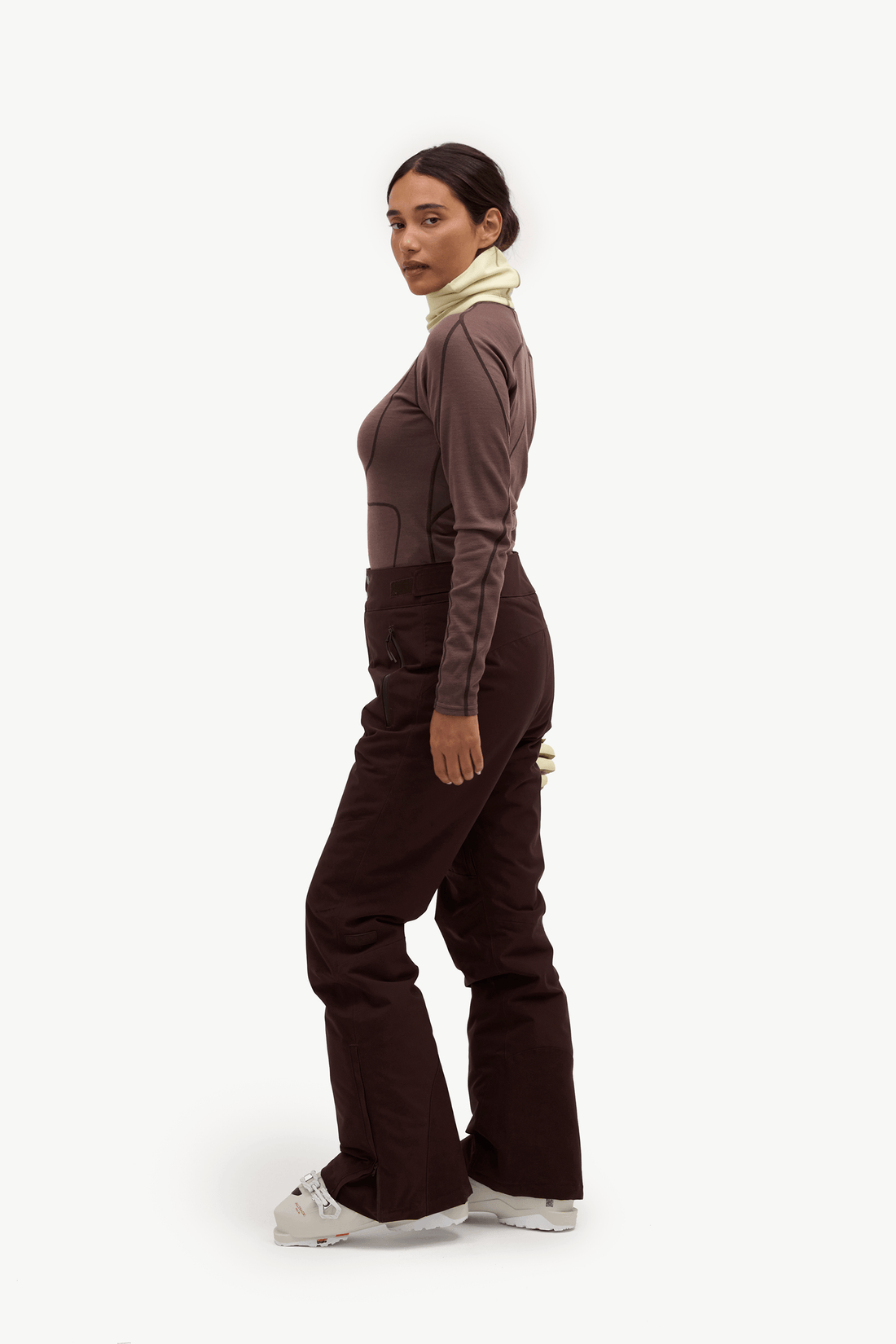 A woman modeling a dark brown long-sleeve top and matching wide-leg brown pants, styled with a cream turtleneck and white sneakers, shown in profile against a plain white studio background.