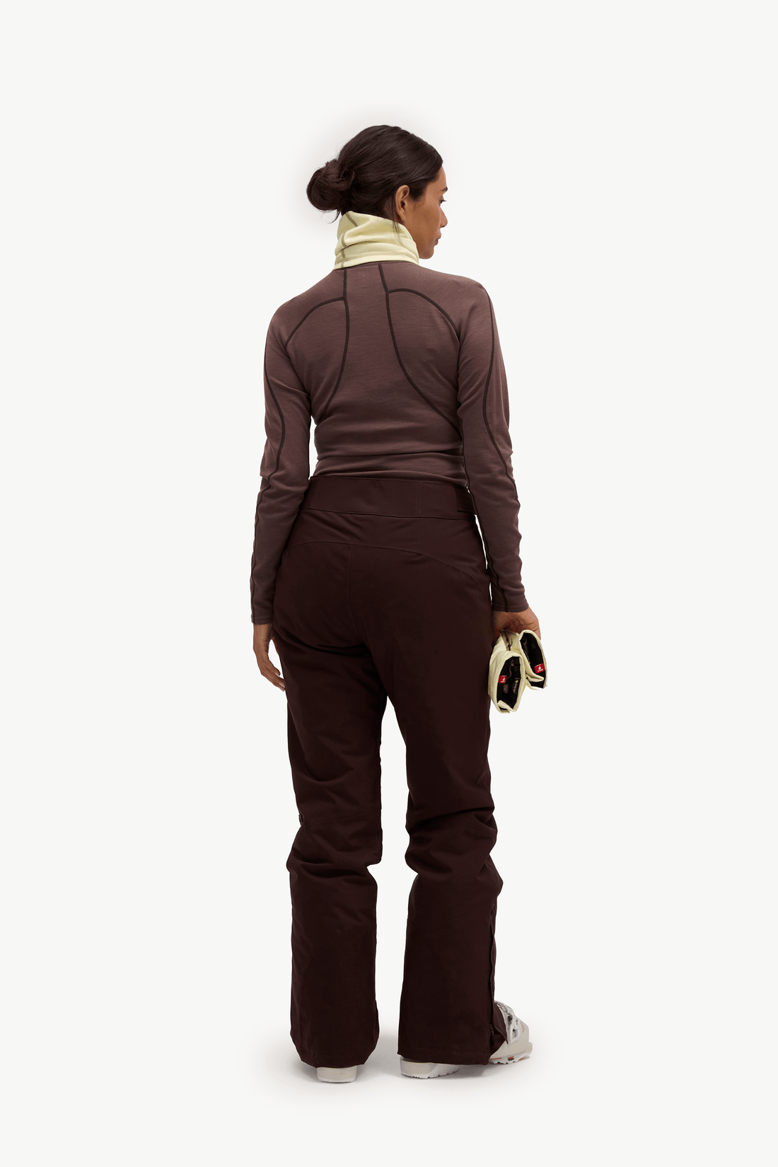 A woman shown from the back wearing a dark brown long-sleeve athletic top and matching dark brown pants, with a light beige neck gaiter and white sneakers, against a plain white studio background.