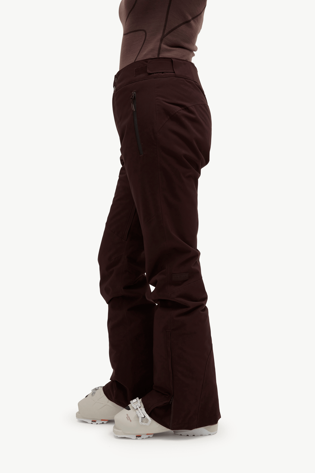 Side view of a person wearing dark brown ski pants with a zippered pocket and light-colored ski boots, shown against a plain light background.