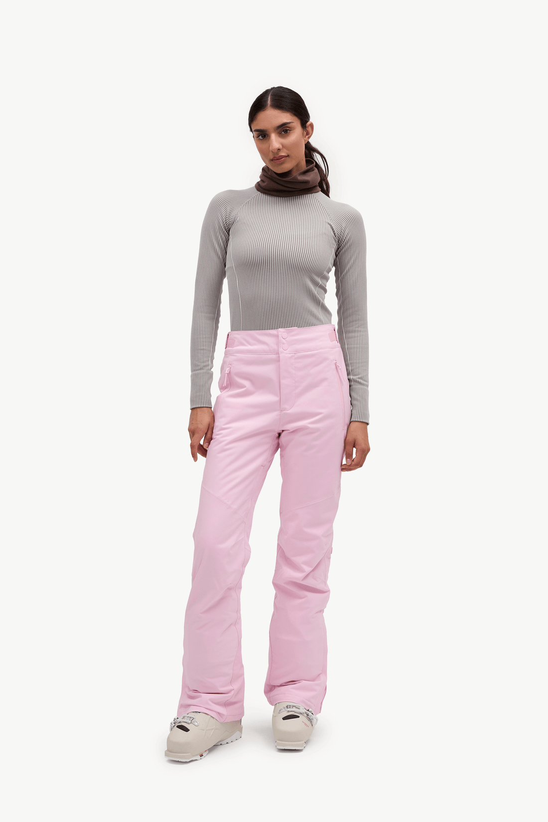 A woman modeling a pair of light pink ski pants with a gray long-sleeve top and brown neck gaiter, standing against a solid off-white background.