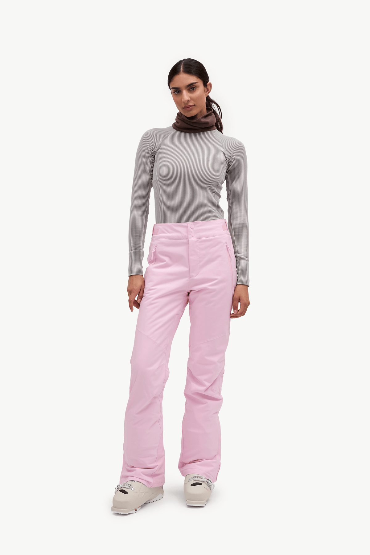 A woman modeling a pair of light pink ski pants with a gray long-sleeve top and brown neck gaiter, standing against a solid off-white background.