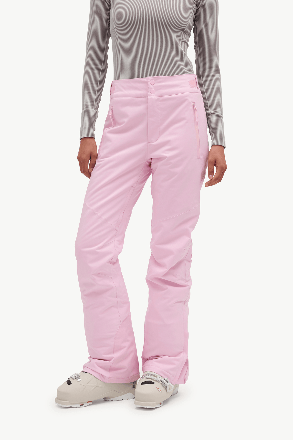 A model wearing light pink ski pants, a gray ribbed long-sleeve top, and ski boots against a plain white studio background.