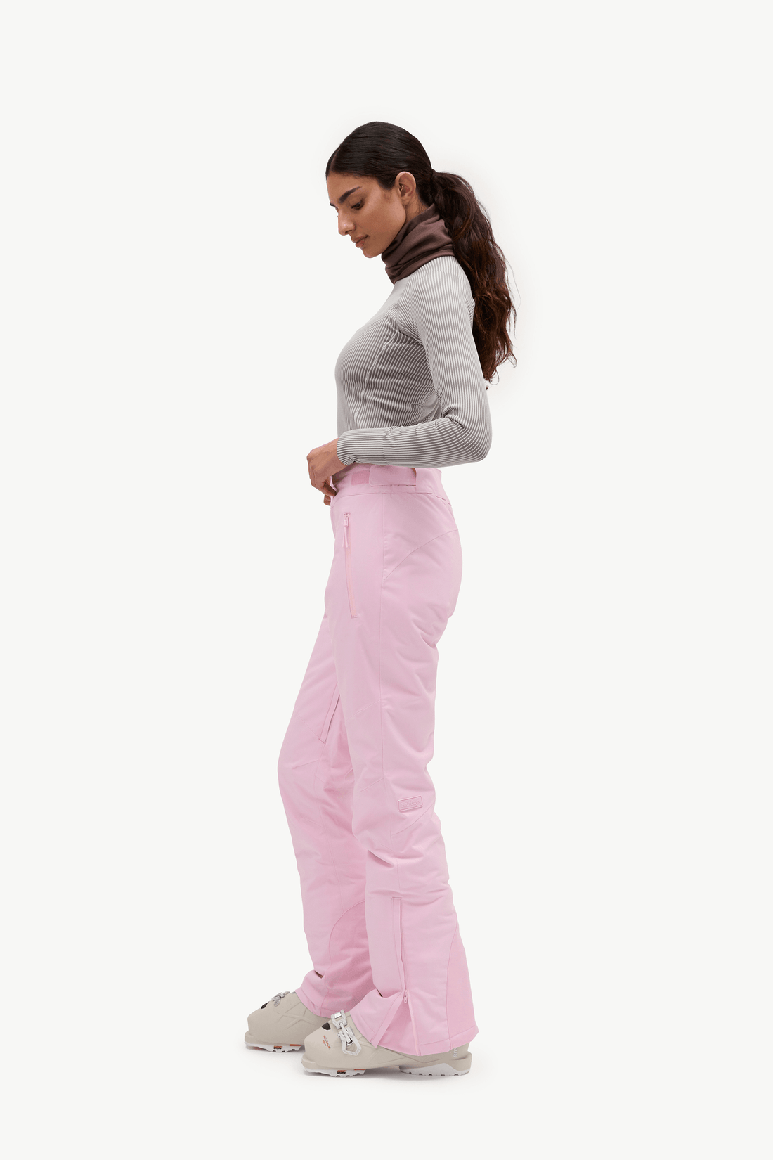 A woman in a side profile wearing light pink ski pants, a gray long-sleeve top, and light-colored sneakers, set against a plain white studio background.