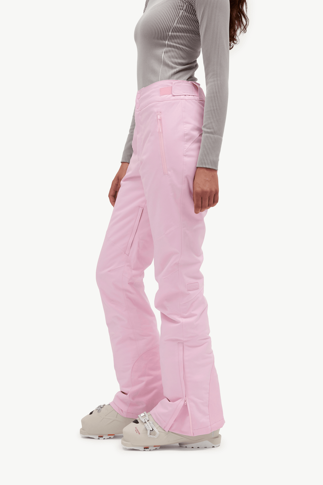 A side profile view of a model wearing light pink ski pants, a gray ribbed long-sleeve top, and beige ski boots against a plain white studio background.