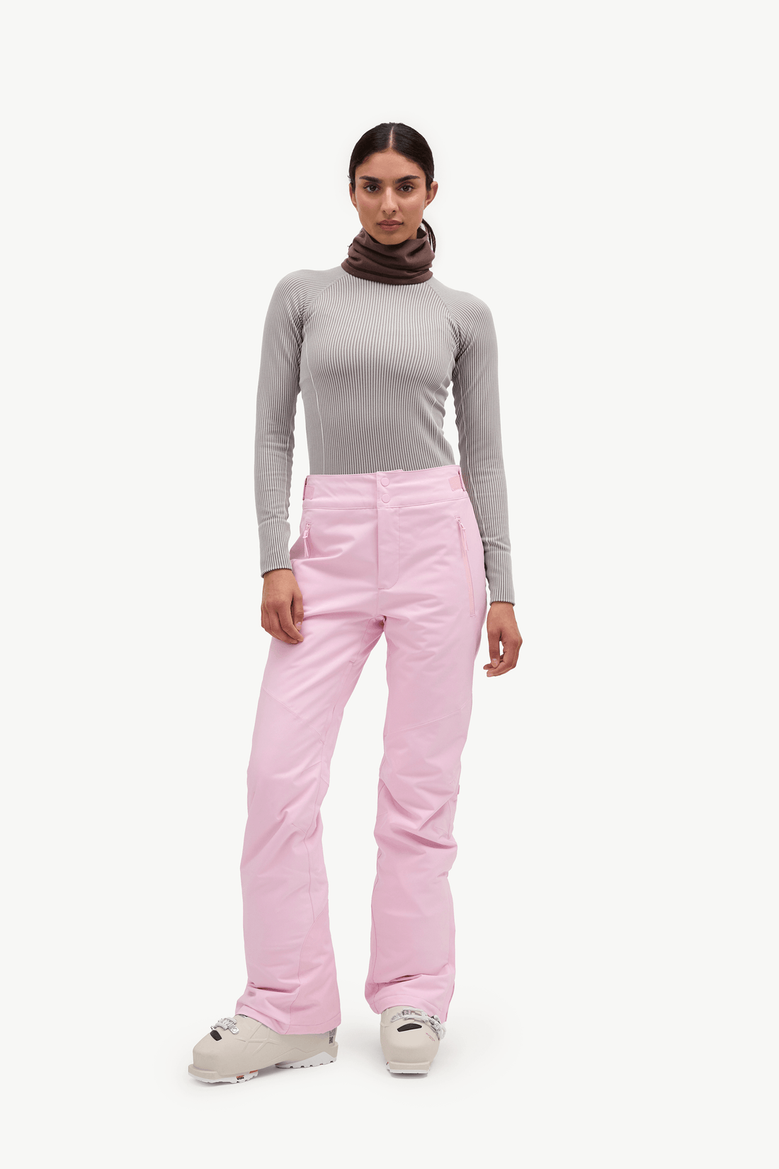 A model stands against a plain white studio background wearing light pink ski pants, a gray long-sleeve top, a dark brown neck warmer, and white ski boots.