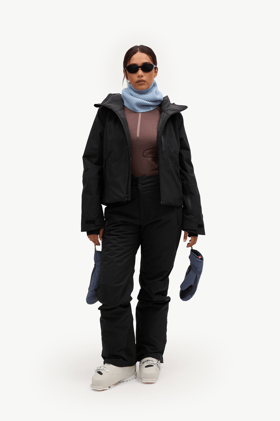 A model stands against a plain white studio background wearing a black ski jacket, black ski pants, a light blue scarf, a brown top, and white sneakers, while holding a pair of dark blue gloves.