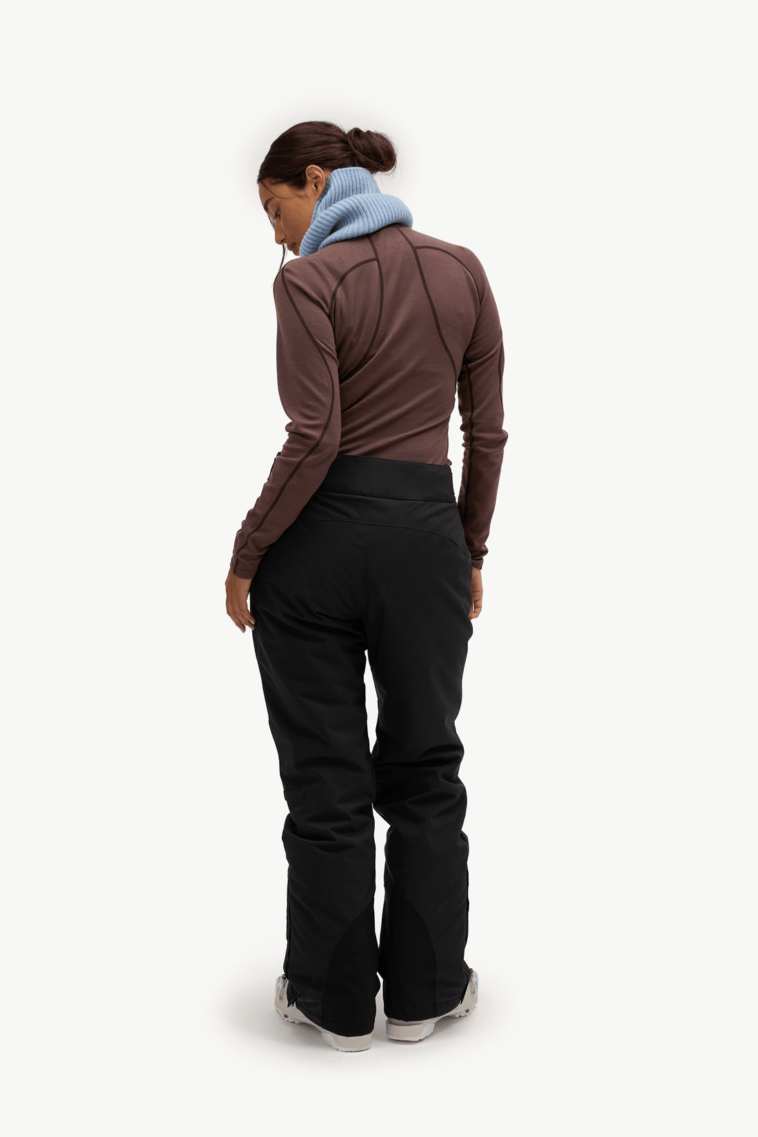 A model stands against a plain white studio background wearing a black ski jacket, black ski pants, a light blue scarf, a brown top, and white sneakers, while holding a pair of dark blue gloves.