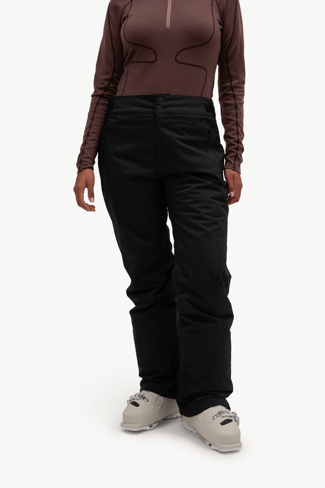 A model wearing black ski pants, a brown long-sleeve athletic top, and light gray ski boots, shown from the chest down against a plain white studio background.