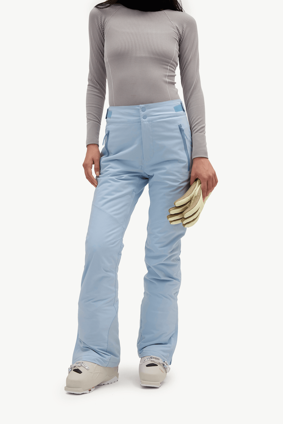 A model wearing light blue ski pants, a gray ribbed long-sleeve top, and light-colored ski boots, holding a pair of gloves against a plain white studio background.