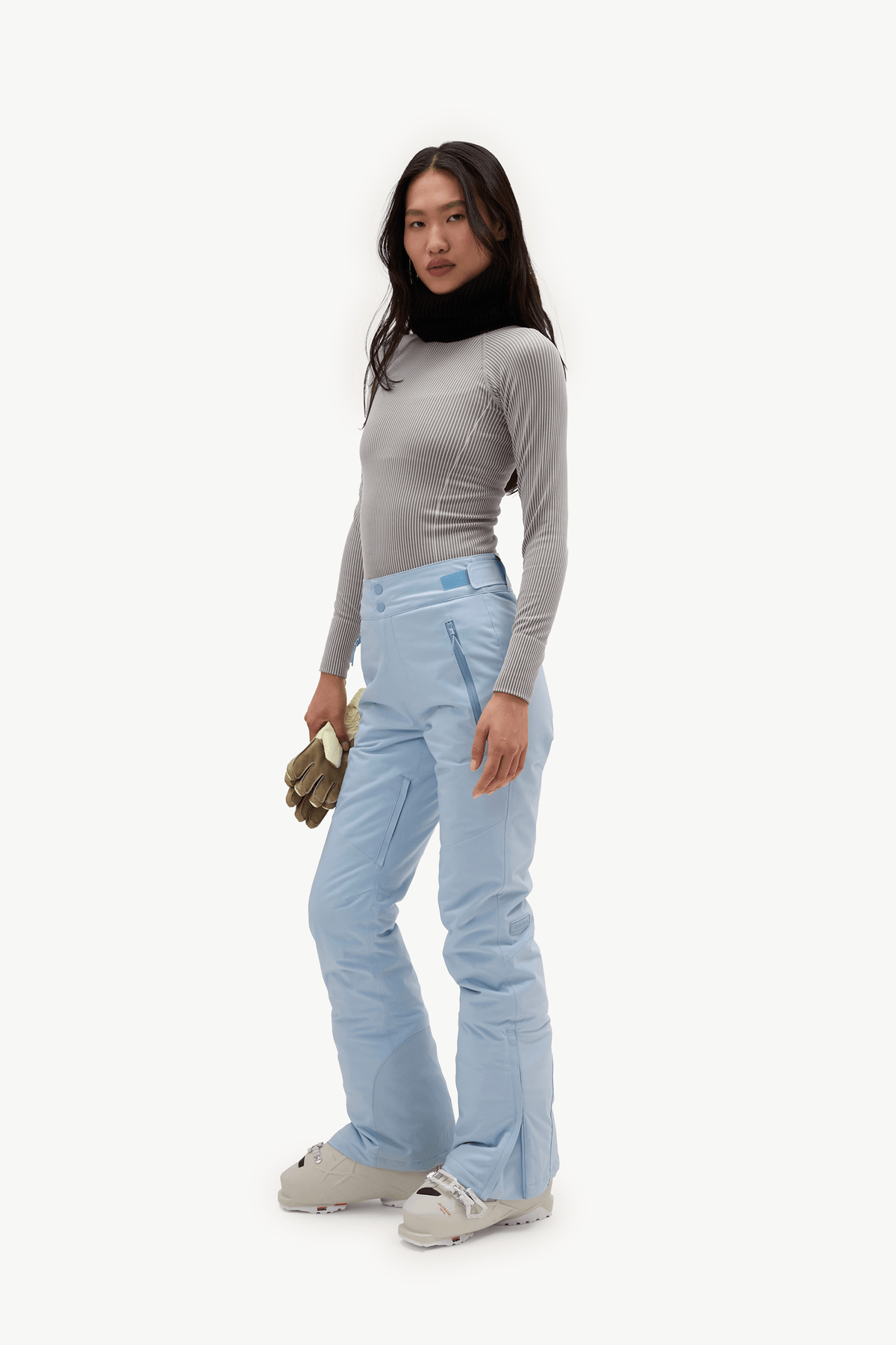 A model stands in profile wearing a light gray long-sleeve top, light blue ski pants, and light gray ski boots, holding a small olive green pouch against a plain white studio background.