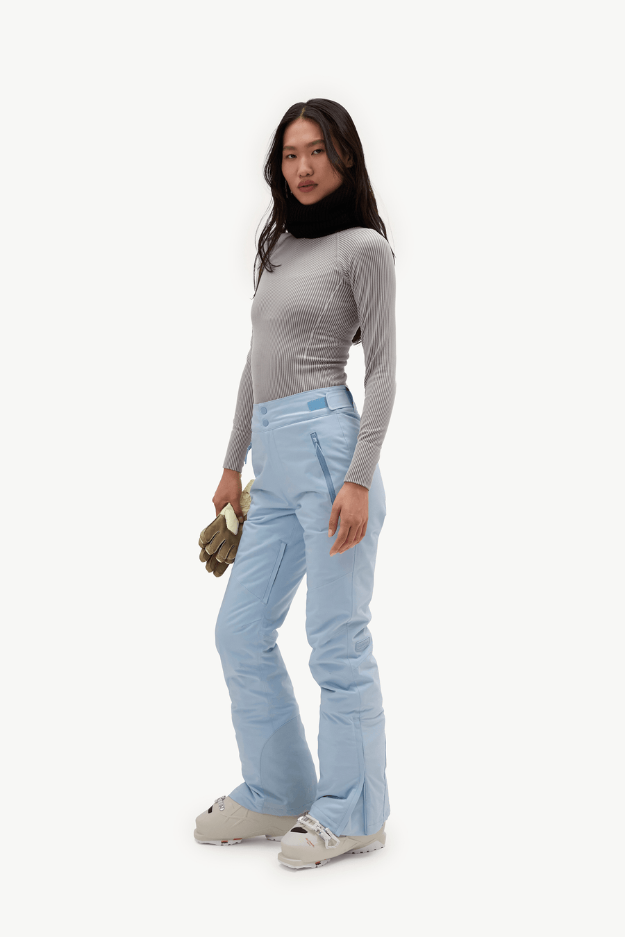 A model stands in profile wearing a light gray long-sleeve top, light blue ski pants, and light gray ski boots, holding a small olive green pouch against a plain white studio background.