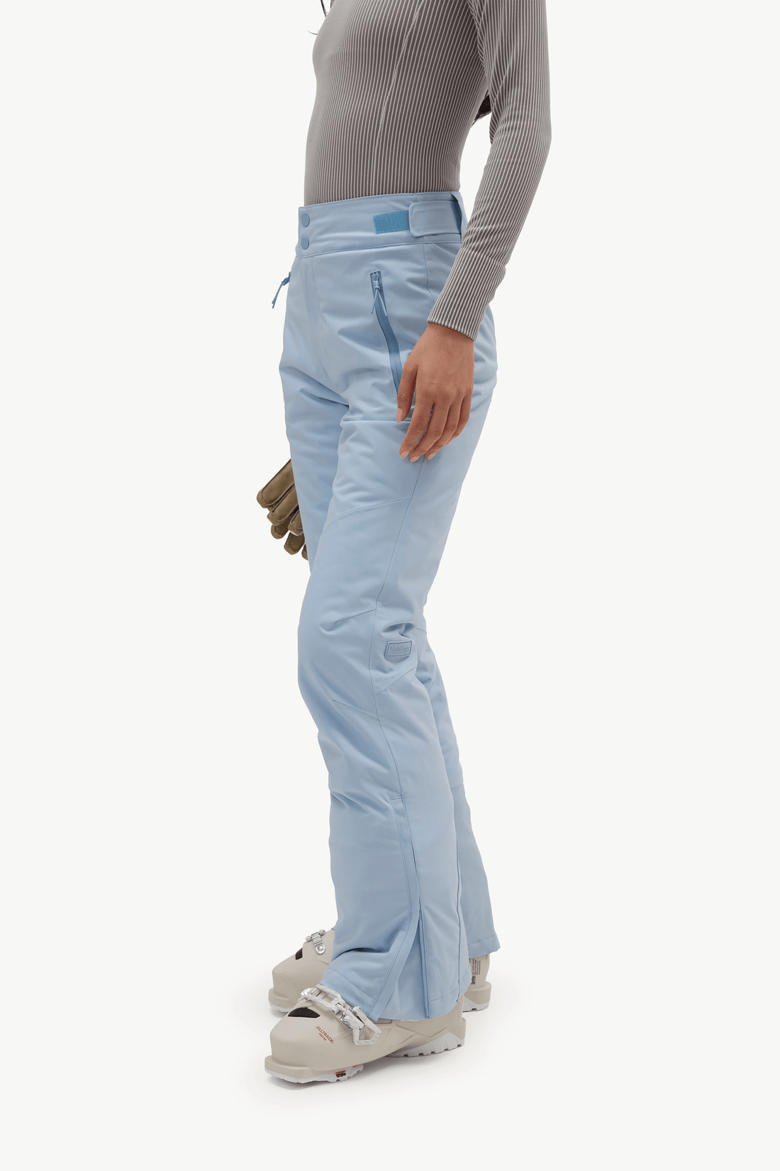 A model stands in profile wearing a light gray long-sleeve top, light blue ski pants, and light gray ski boots, holding a small olive green pouch against a plain white studio background.