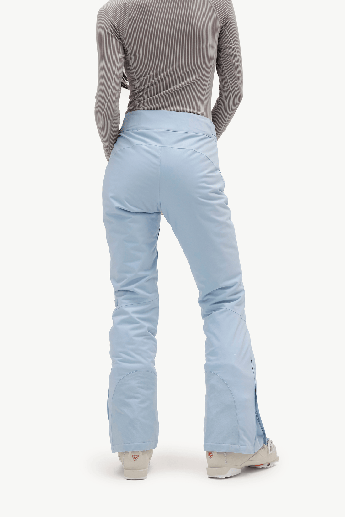 A model stands in profile wearing a light gray long-sleeve top, light blue ski pants, and light gray ski boots, holding a small olive green pouch against a plain white studio background.