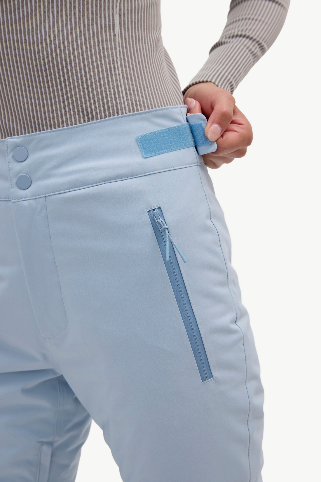 Close-up of a person wearing light blue ski pants, showing the waistband with a hook-and-loop closure being adjusted by a hand, alongside a zippered pocket.