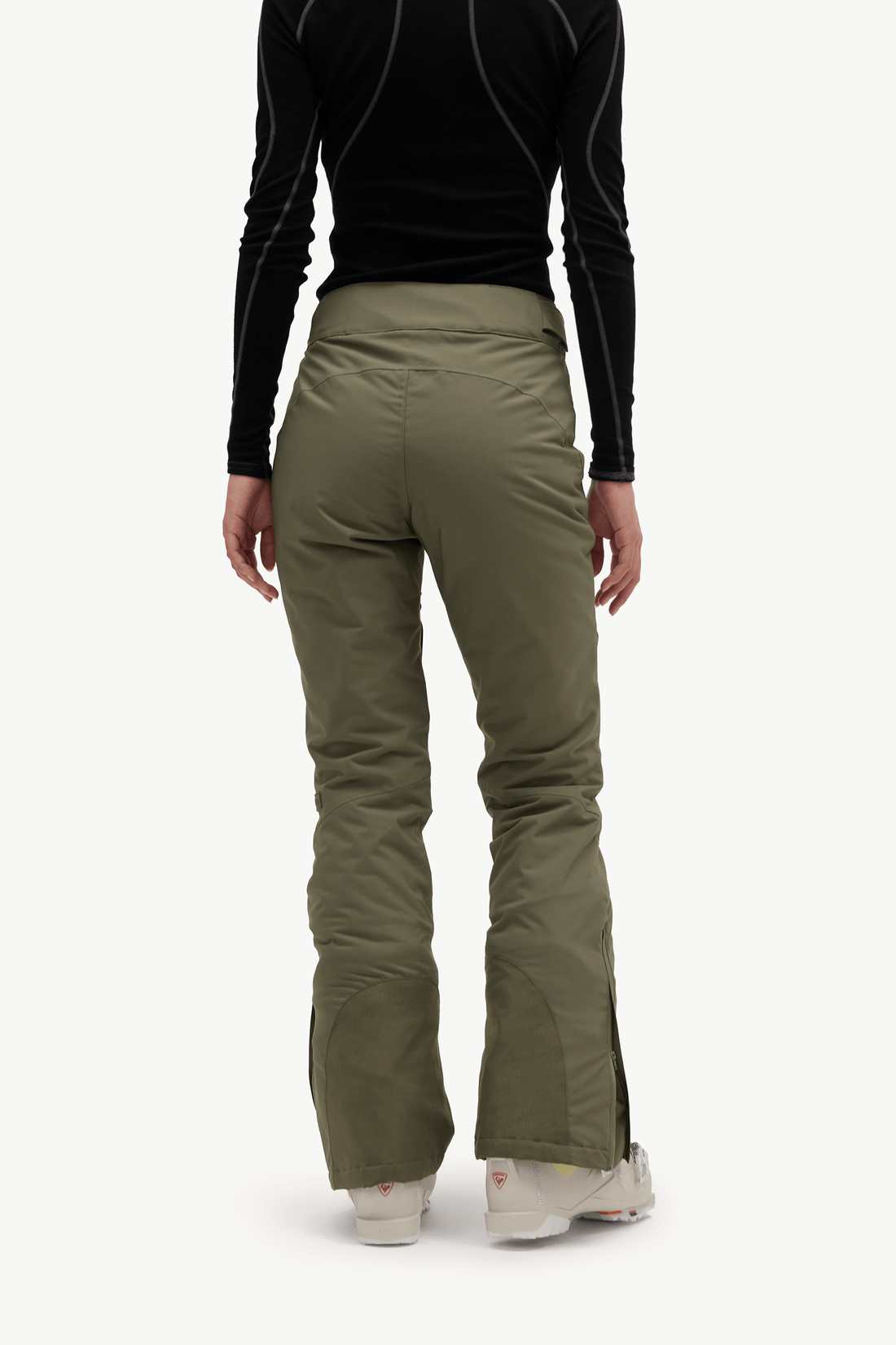A person shown from the back wearing olive green ski pants, a black long-sleeve top, and white sneakers against a plain light background.
