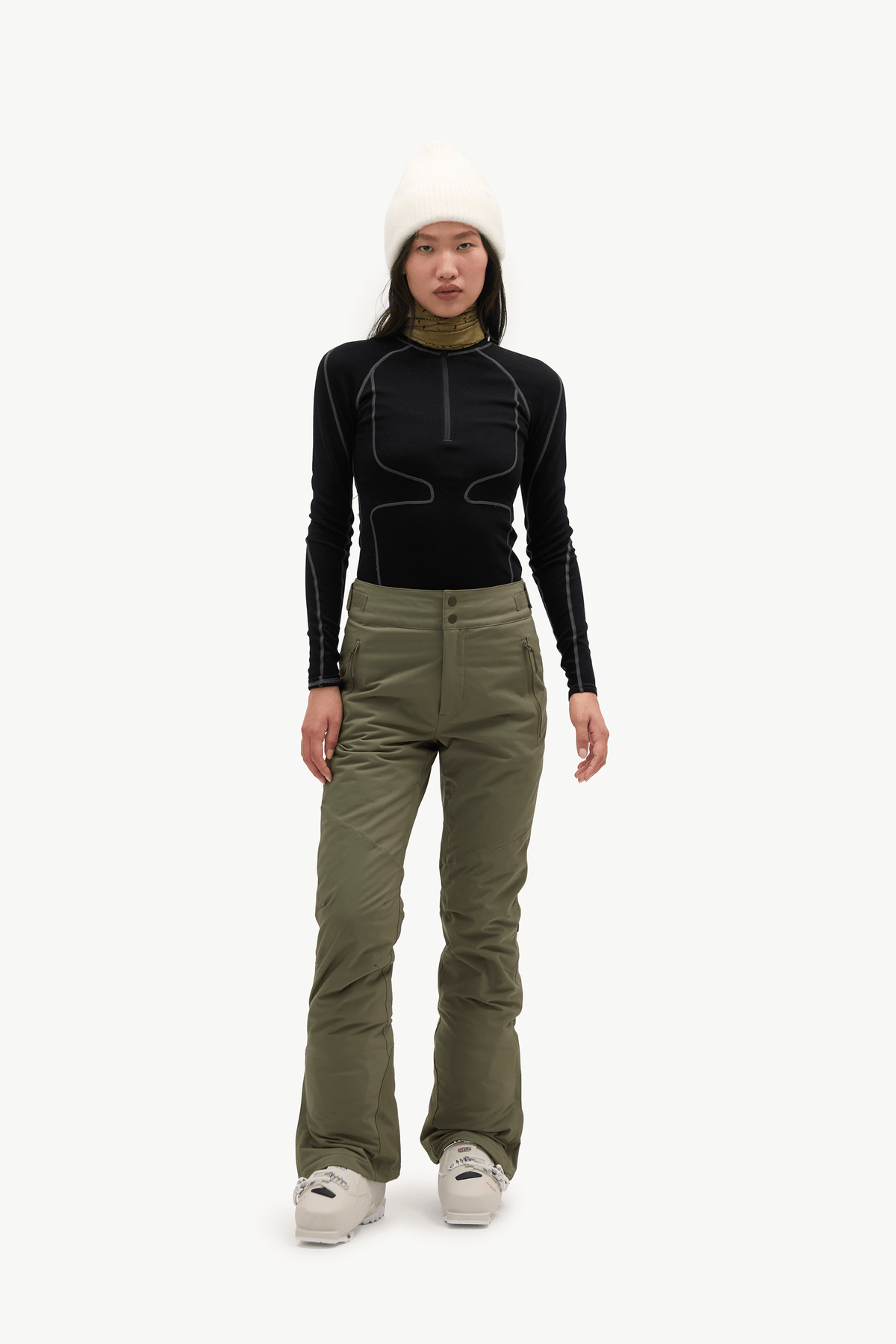 A model stands against a light gray studio background wearing a black long-sleeve athletic top with contrast stitching, olive green ski pants, a white beanie, and white sneakers.