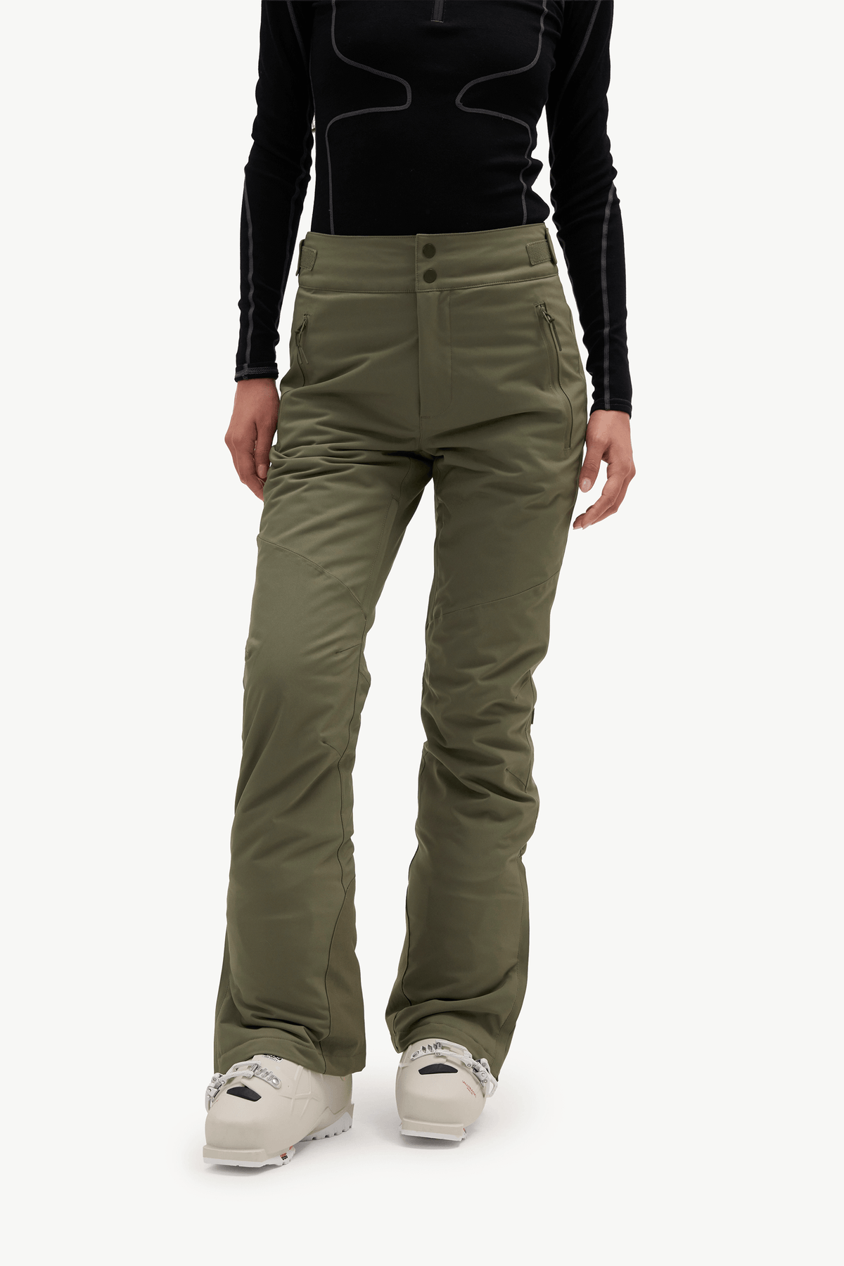 A model stands against a light gray studio background wearing a black long-sleeve athletic top with contrast stitching, olive green ski pants, a white beanie, and white sneakers.