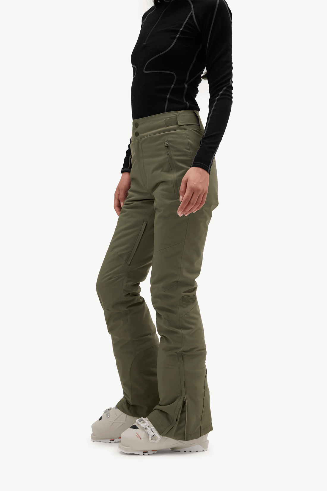 A model stands against a light gray studio background wearing a black long-sleeve athletic top with contrast stitching, olive green ski pants, a white beanie, and white sneakers.