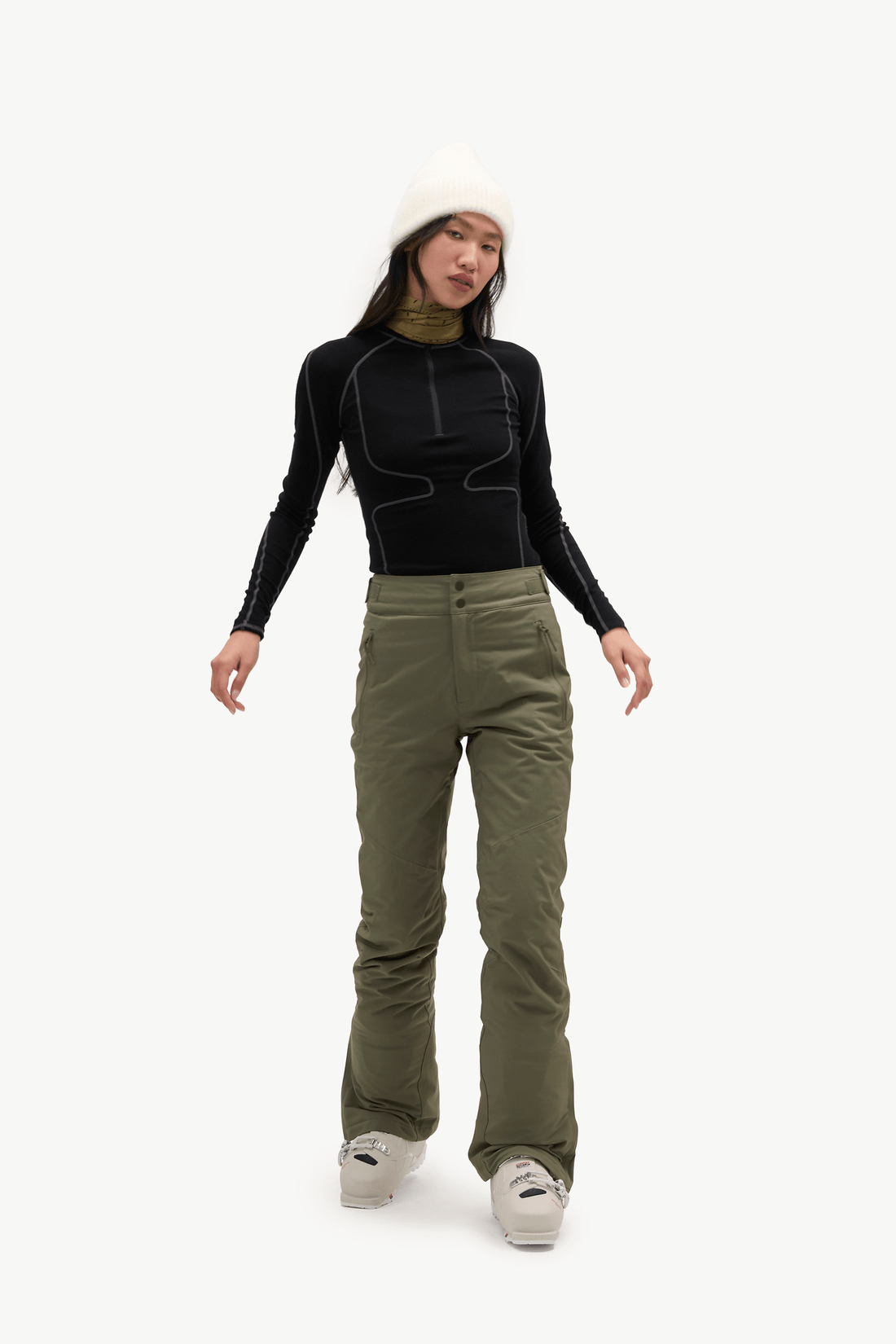 A model stands against a light gray studio background wearing a black long-sleeve athletic top with contrast stitching, olive green ski pants, a white beanie, and white sneakers.