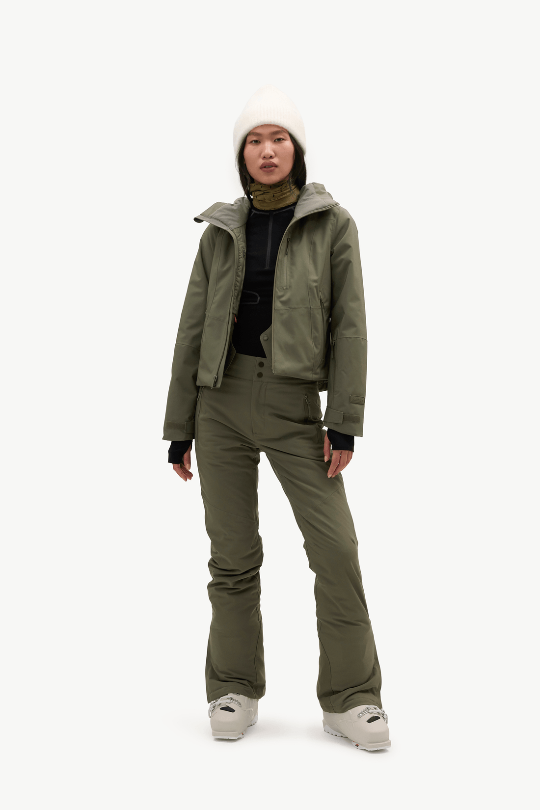 A model stands against a plain white studio background wearing an olive green ski jacket, matching ski pants, a black turtleneck, a white knit beanie, and light gray sneakers.