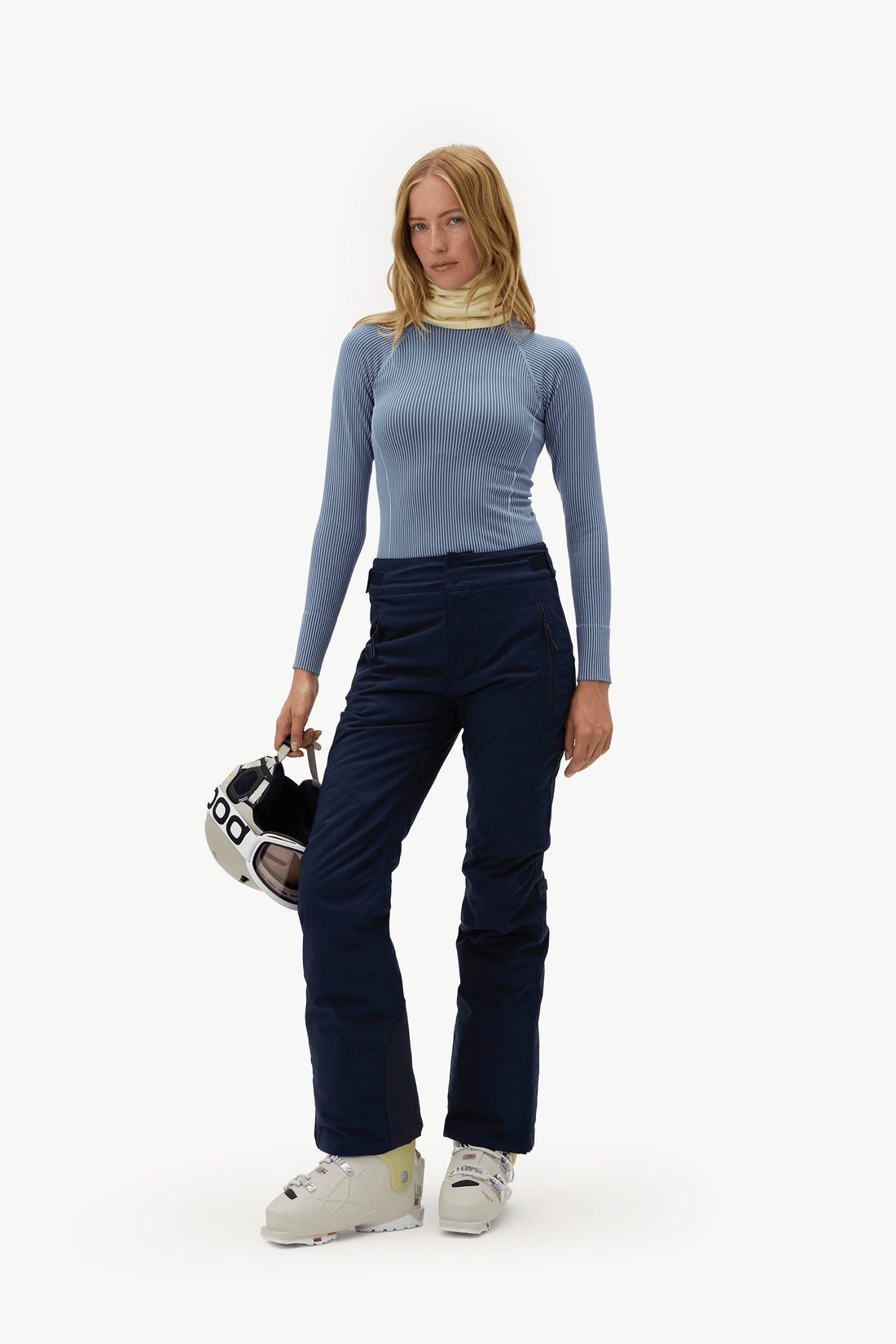 A woman modeling a light blue ribbed long-sleeve top and dark navy ski pants, holding a ski helmet against a clean white studio background.