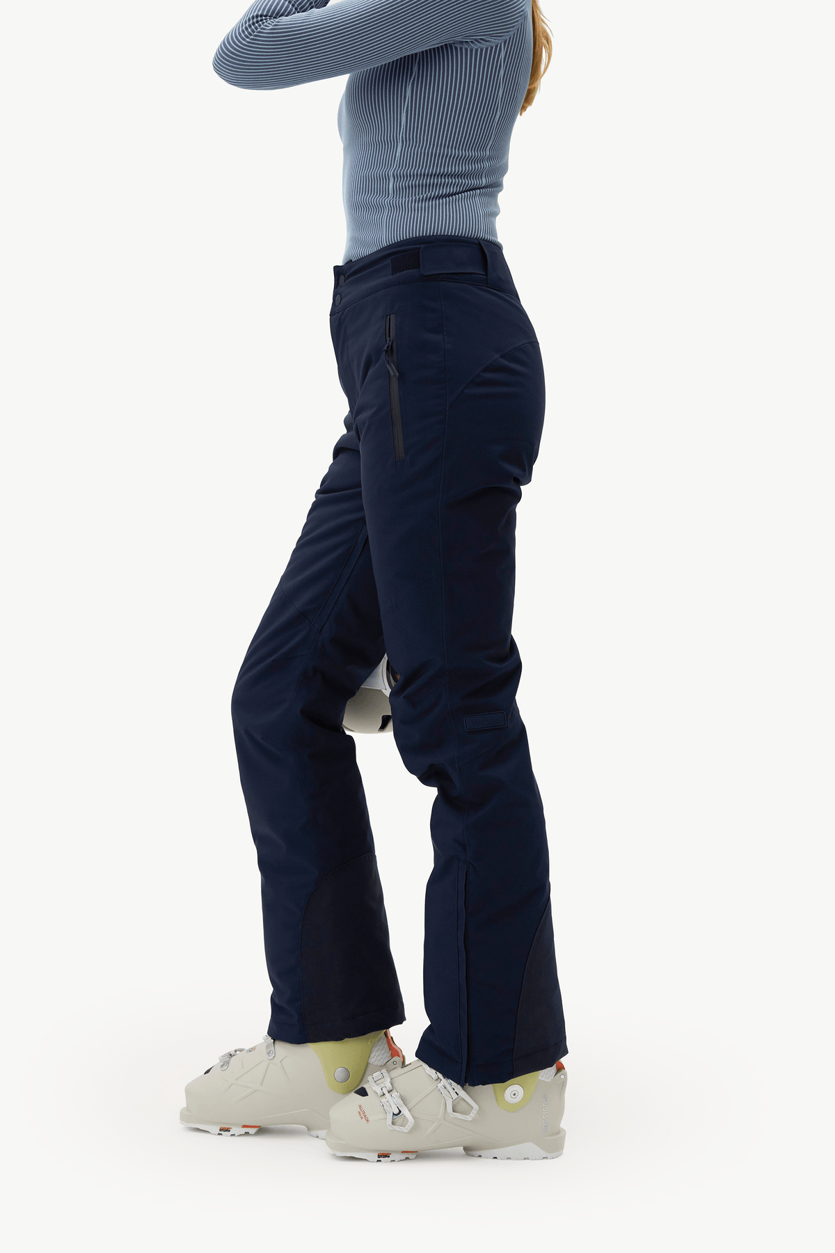 A side profile of a model wearing dark navy ski pants, a light blue ribbed long-sleeve top, and off-white ski boots against a plain white studio background.