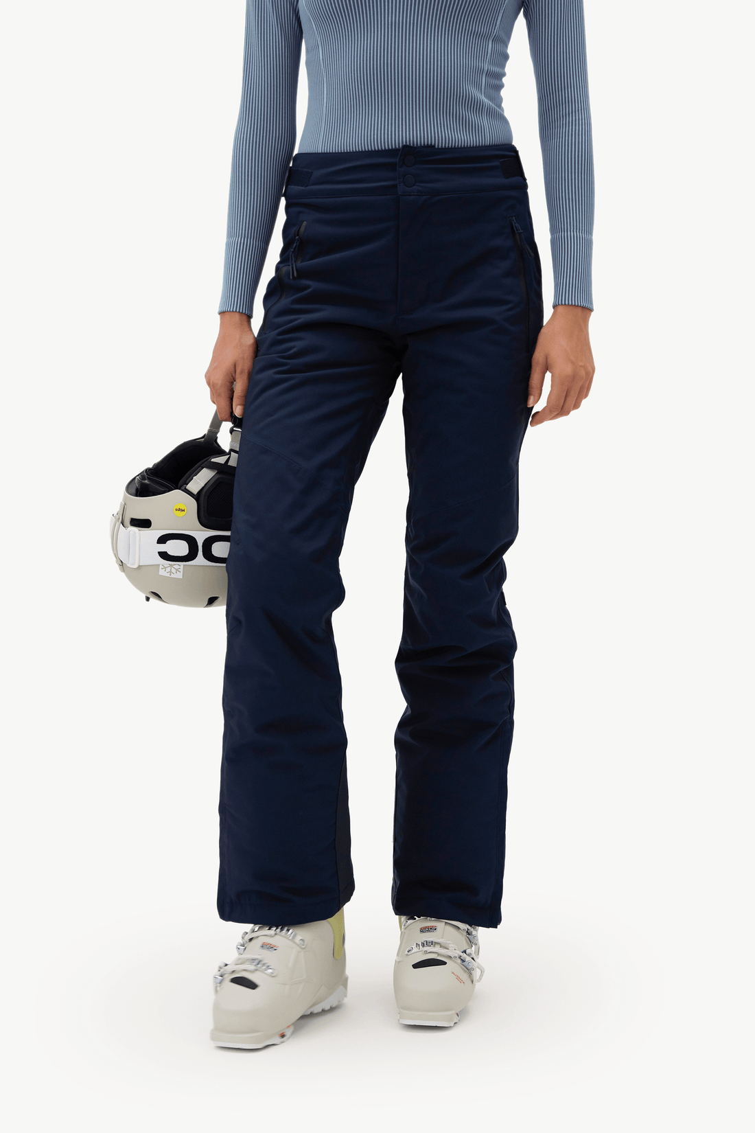 A model wearing dark navy ski pants, a light blue ribbed long-sleeve top, and white ski boots, holding a white and black POC ski helmet against a clean white background.