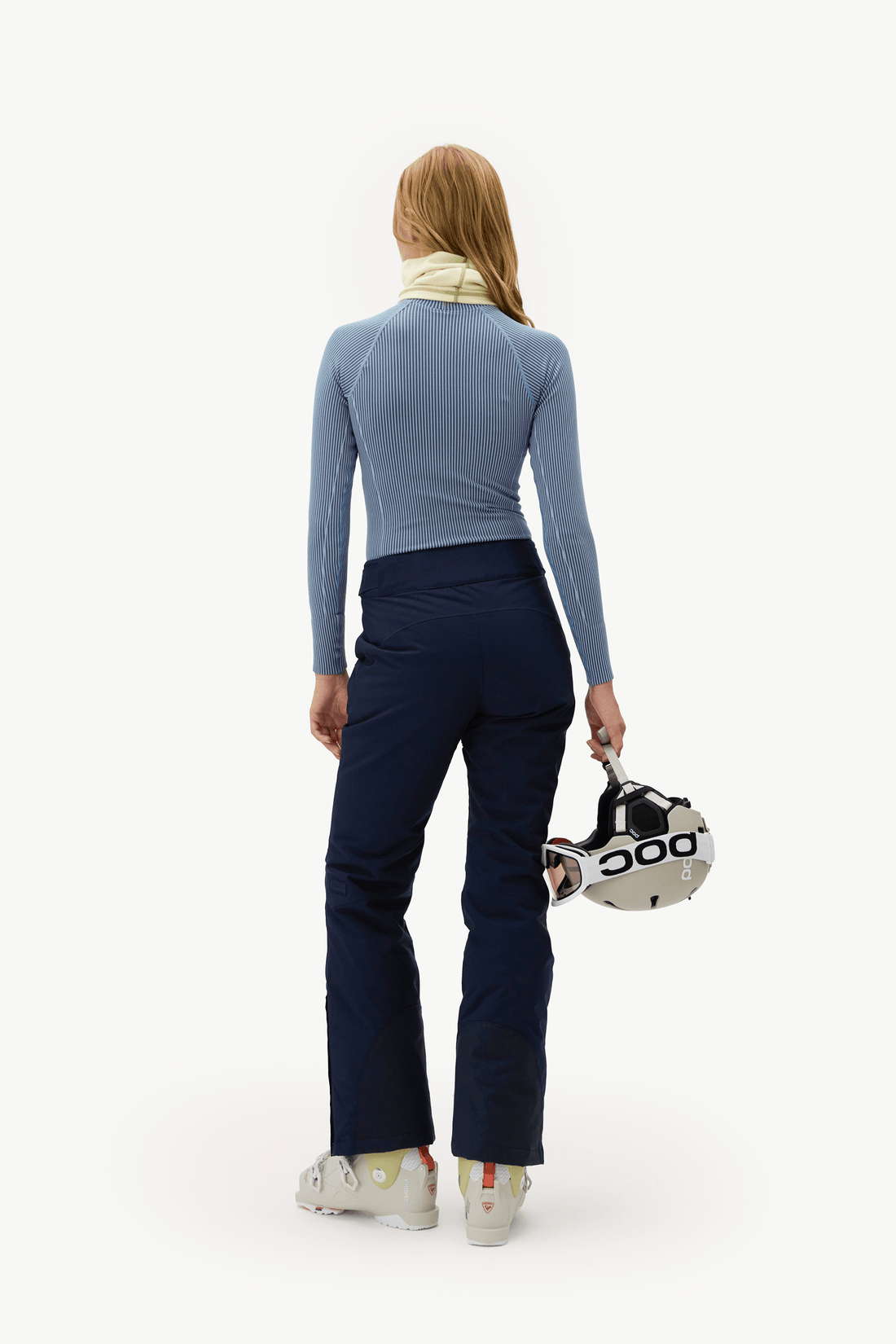 A woman is shown from behind wearing a light blue long-sleeve top, navy blue ski pants, and white ski boots, holding a white and black ski helmet against a plain white studio background.