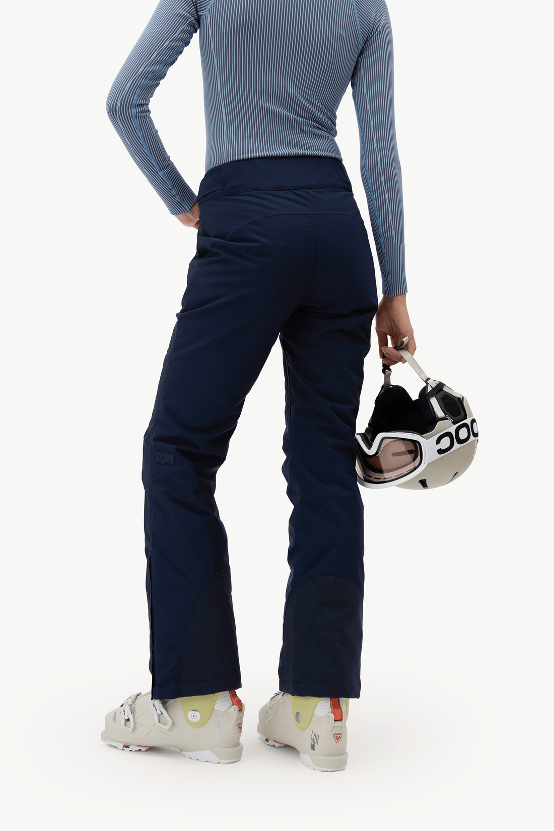 A model from behind wearing dark navy ski pants, a light blue ribbed long-sleeve top, and white ski boots, holding a black and white ski helmet against a clean white background.