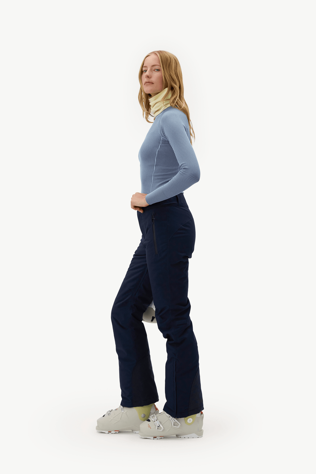 A woman in side profile wearing a light blue long-sleeve top, dark navy ski pants, a light yellow neck gaiter, and white ski boots, standing against a solid white studio background.