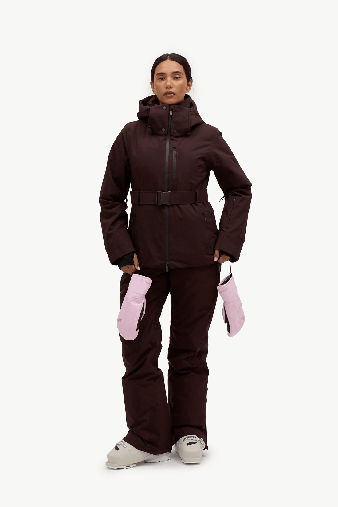 A model stands against a light gray studio background wearing a dark brown ski jacket with a matching belt, dark brown ski pants, light pink ski mittens, and white ski boots.