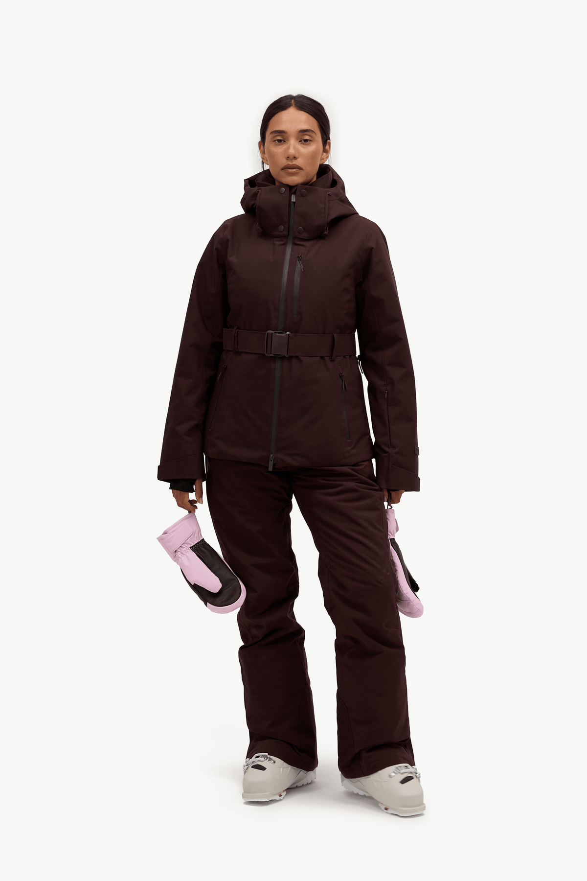 A model stands against a light gray studio background wearing a dark brown ski jacket and matching pants, holding a pair of pink and black gloves.