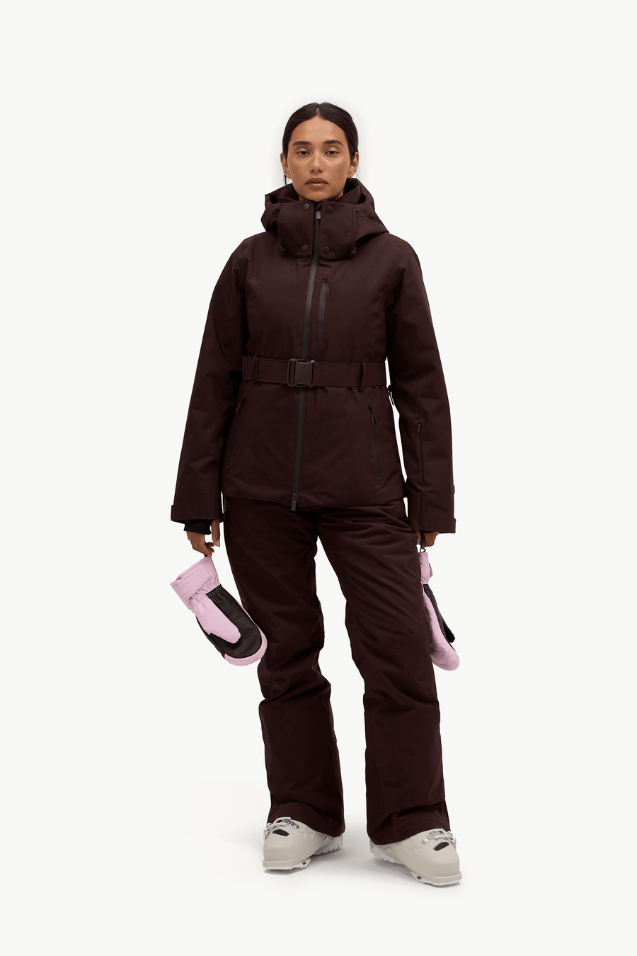 A model stands against a light gray studio background wearing a dark brown ski jacket and matching pants, holding a pair of pink and black gloves.