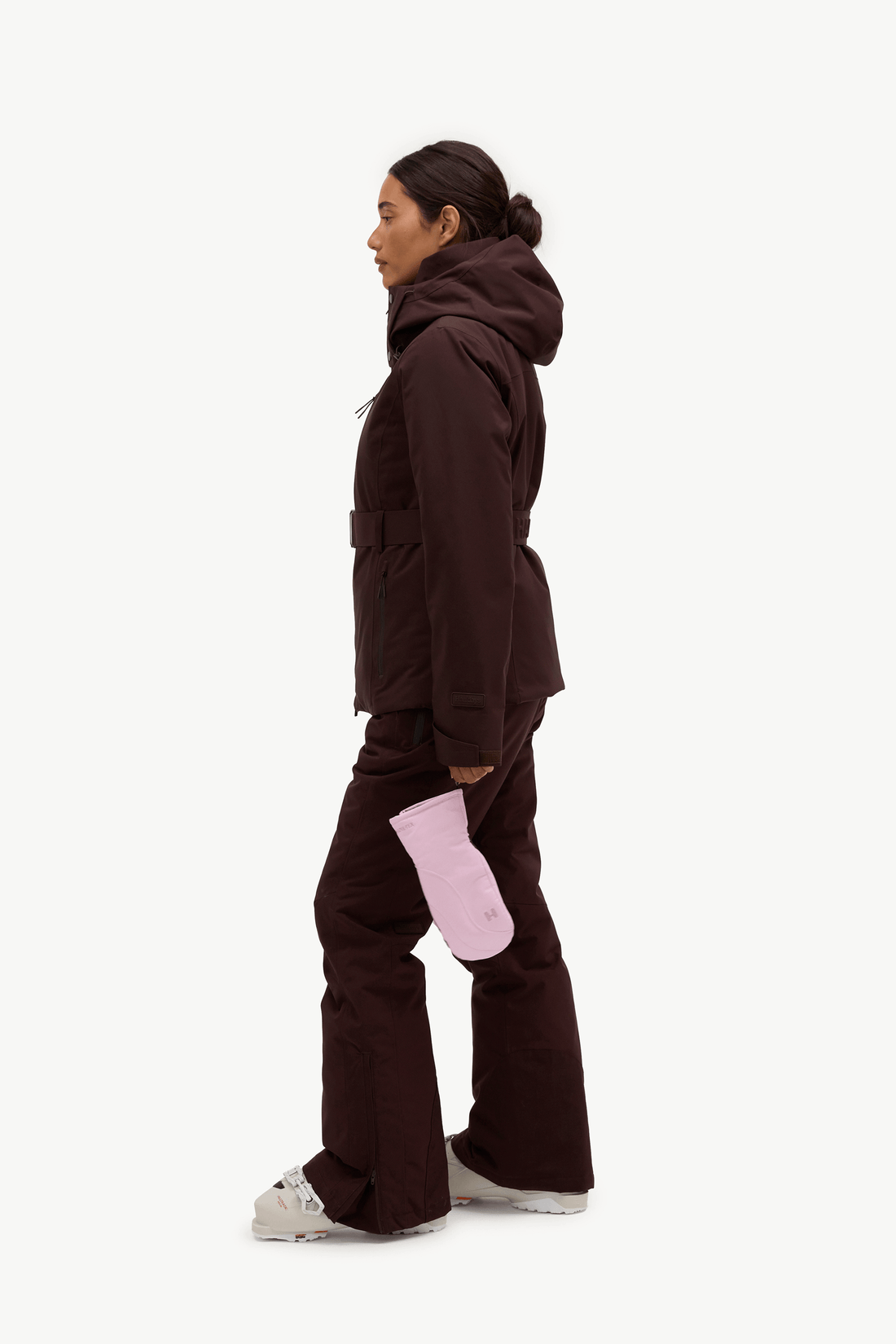 A woman shown in profile wearing a dark brown hooded jacket and matching dark brown pants, with a light pink glove and white sneakers, against a plain light gray studio background.