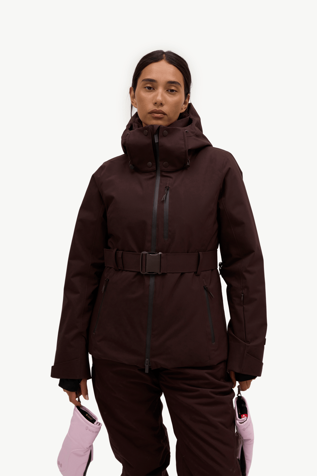 A model wearing a dark brown hooded ski jacket with a matching belt and ski pants, holding light pink ski gloves against a solid white background.