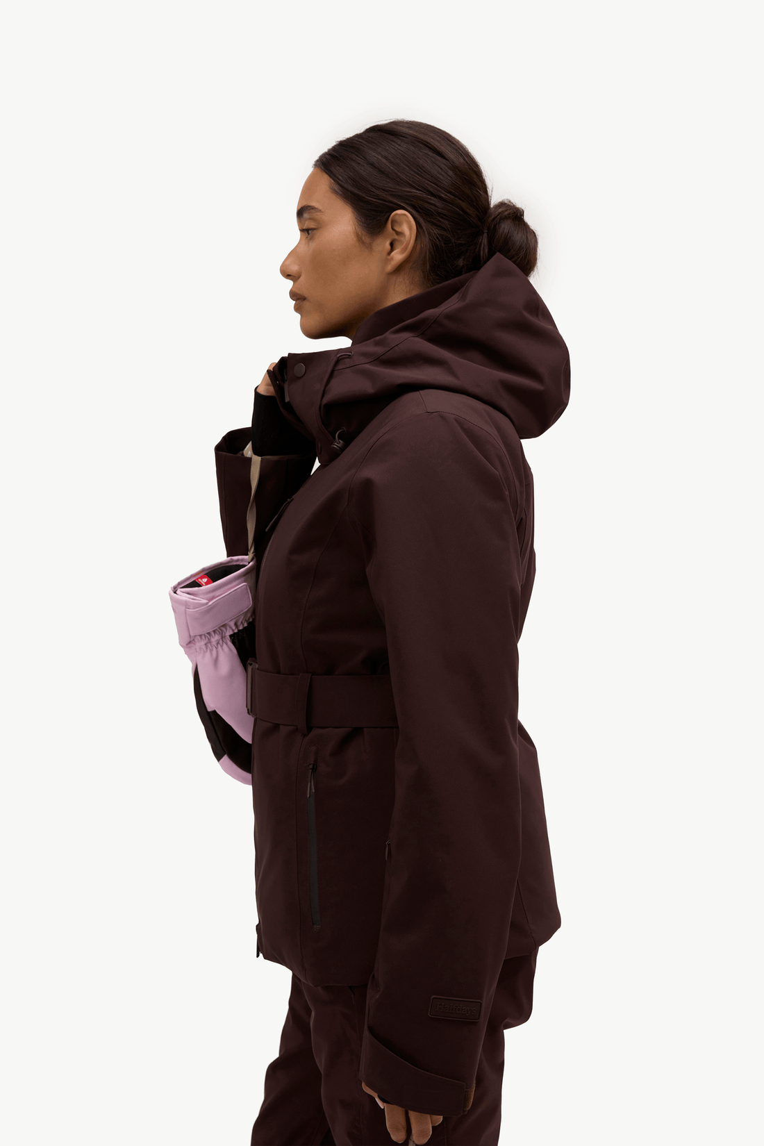 A woman in profile wearing a dark brown hooded jacket and matching pants, with a light pink bag slung over her shoulder, against a plain white studio background.