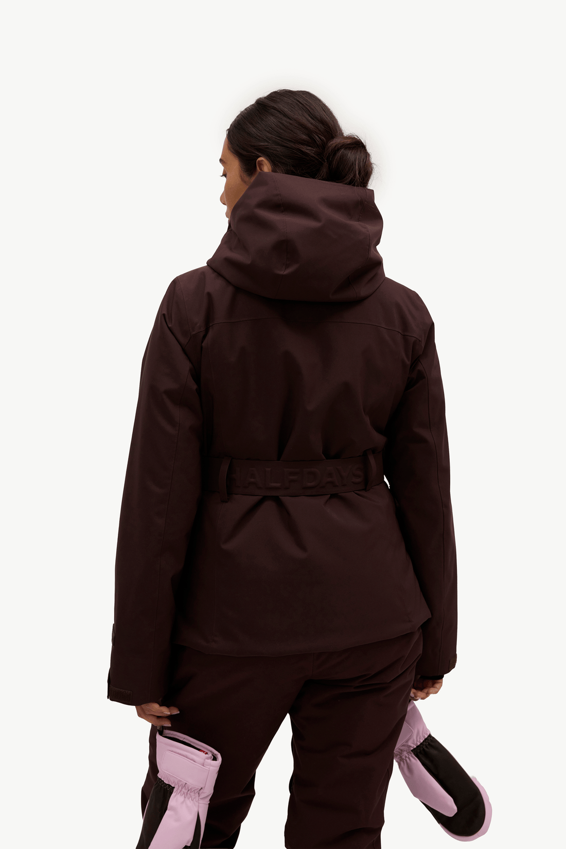 A woman shown from the back wearing a dark brown hooded ski jacket with a matching belt and ski pants, holding pink and black ski boots against a plain white studio background.