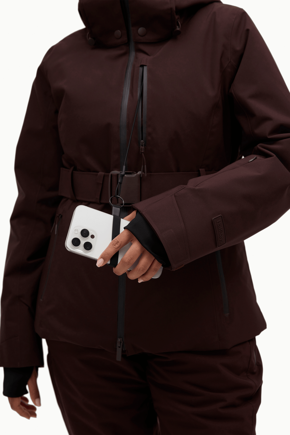 A close-up of a person wearing a dark brown hooded jacket with a front zipper, belt detail, and a white smartphone tucked into a side pocket, against a plain light background.
