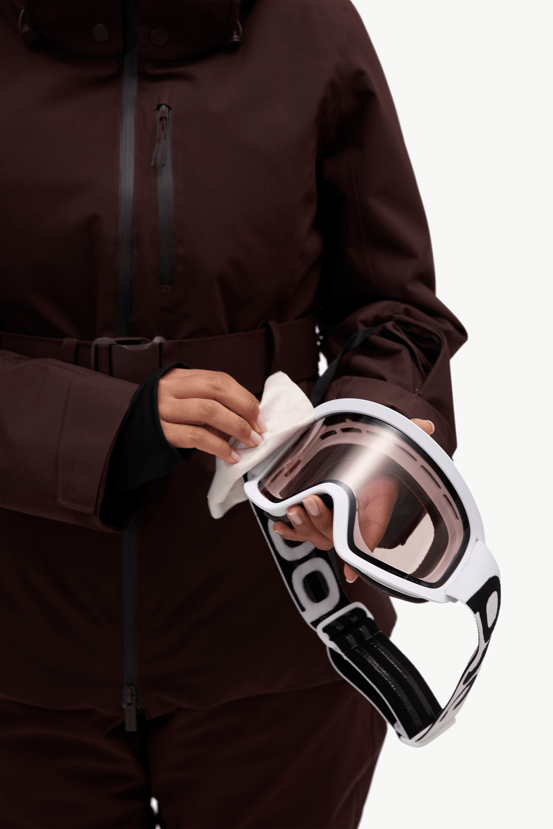 A person in a dark brown ski jacket holds a pair of white ski goggles with a clear lens and black-and-white strap against a clean white background.