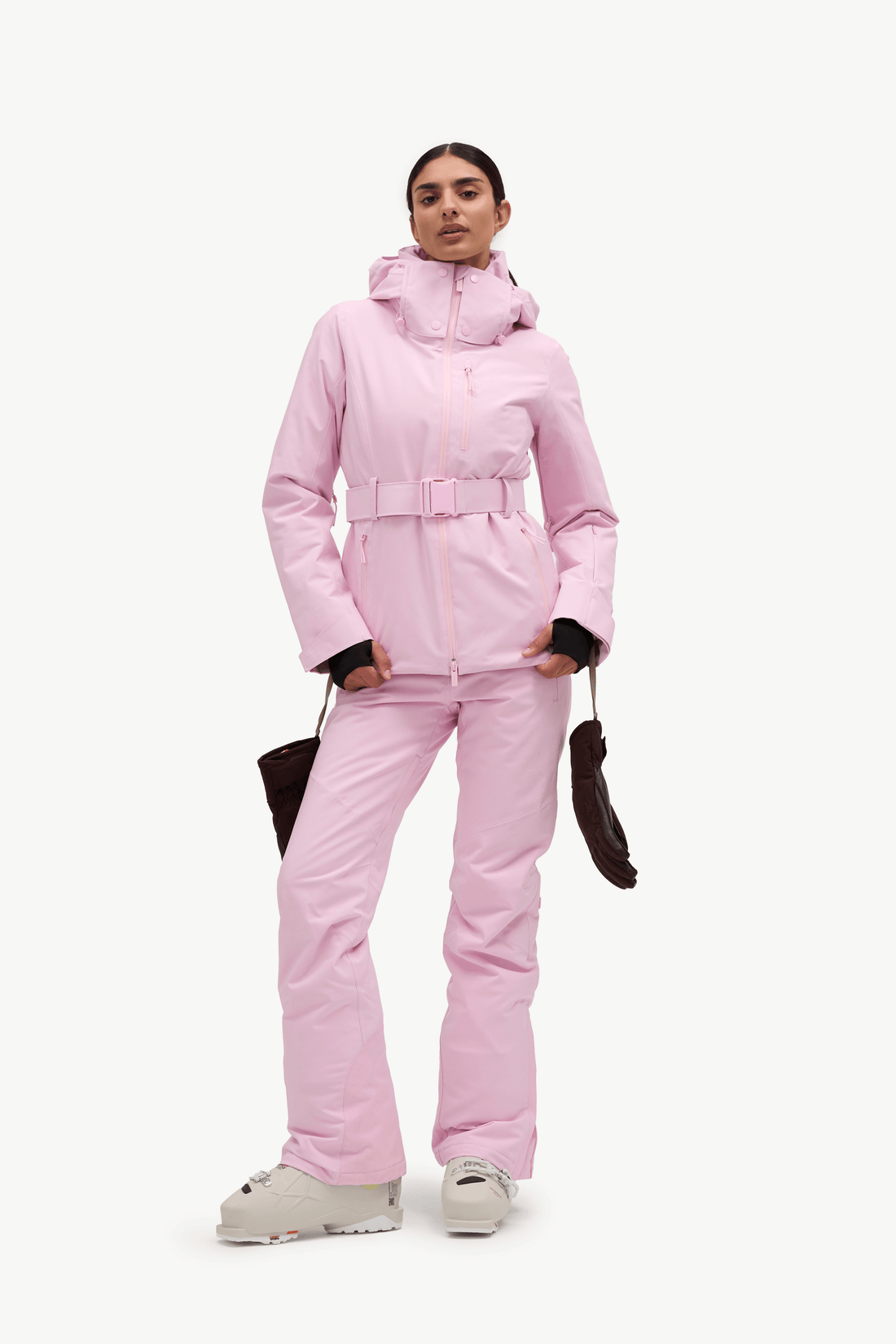A woman in profile wearing a light pink ski jacket and matching ski pants, with dark gloves and white ski boots, against a plain white studio background.