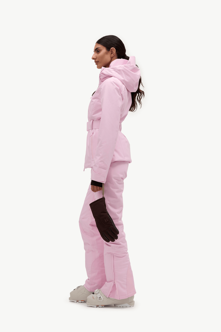 A woman in profile wearing a light pink ski jacket and matching ski pants, with dark gloves and white ski boots, against a plain white studio background.