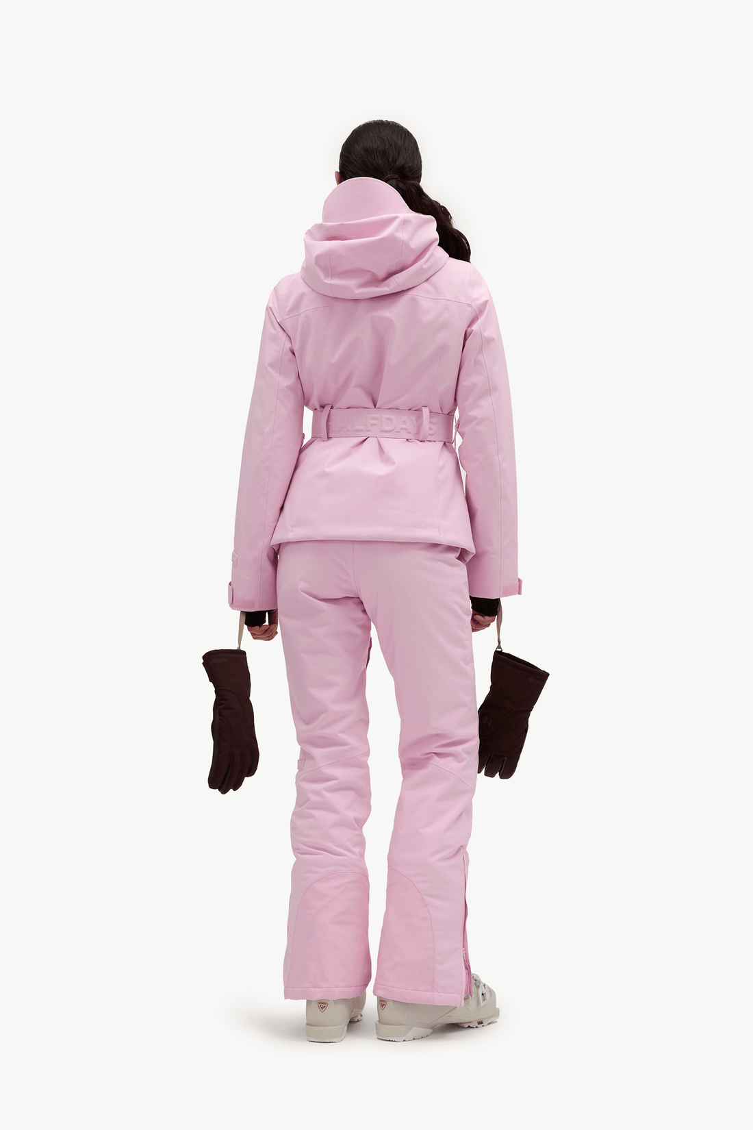 A model shown from the back wearing a light pink hooded ski jacket and matching ski pants, holding black gloves against a plain white studio background.