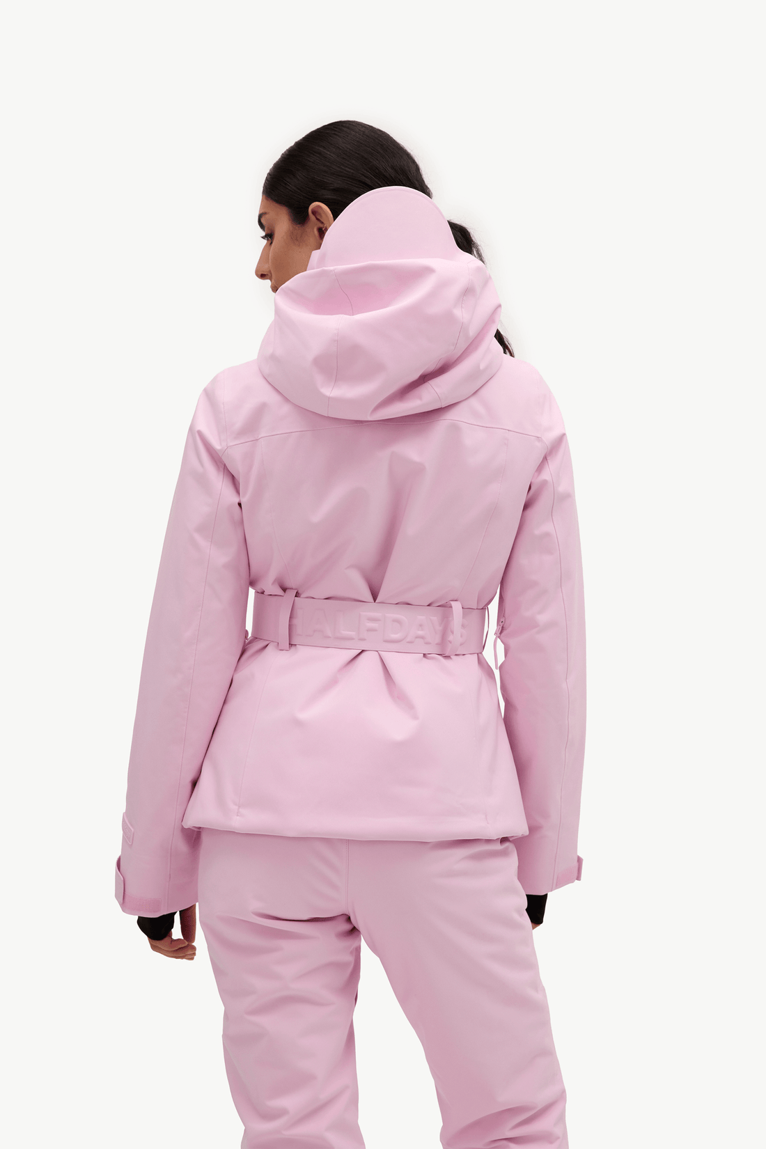 A model shown from the back wearing a light pink hooded ski jacket with a matching belt and matching ski pants against a plain white background.
