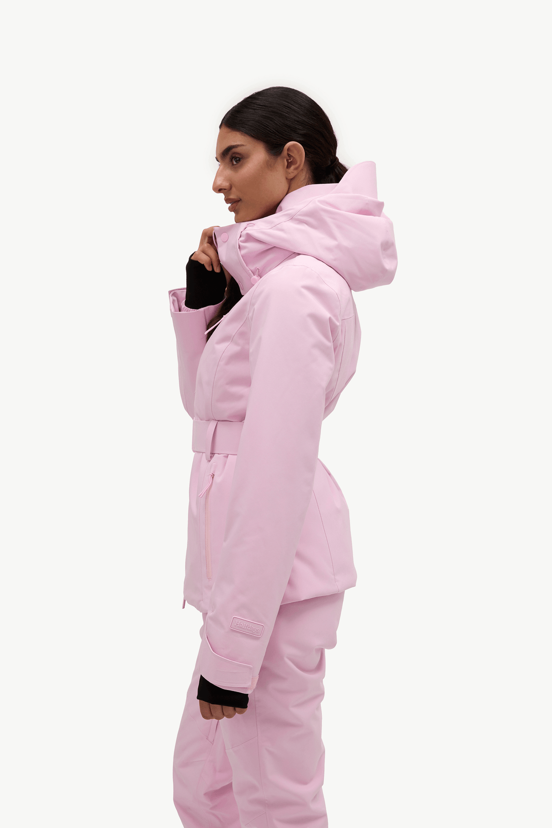 A woman wearing a light pink hooded ski jacket and matching pants, shown in profile against a plain light gray studio background.