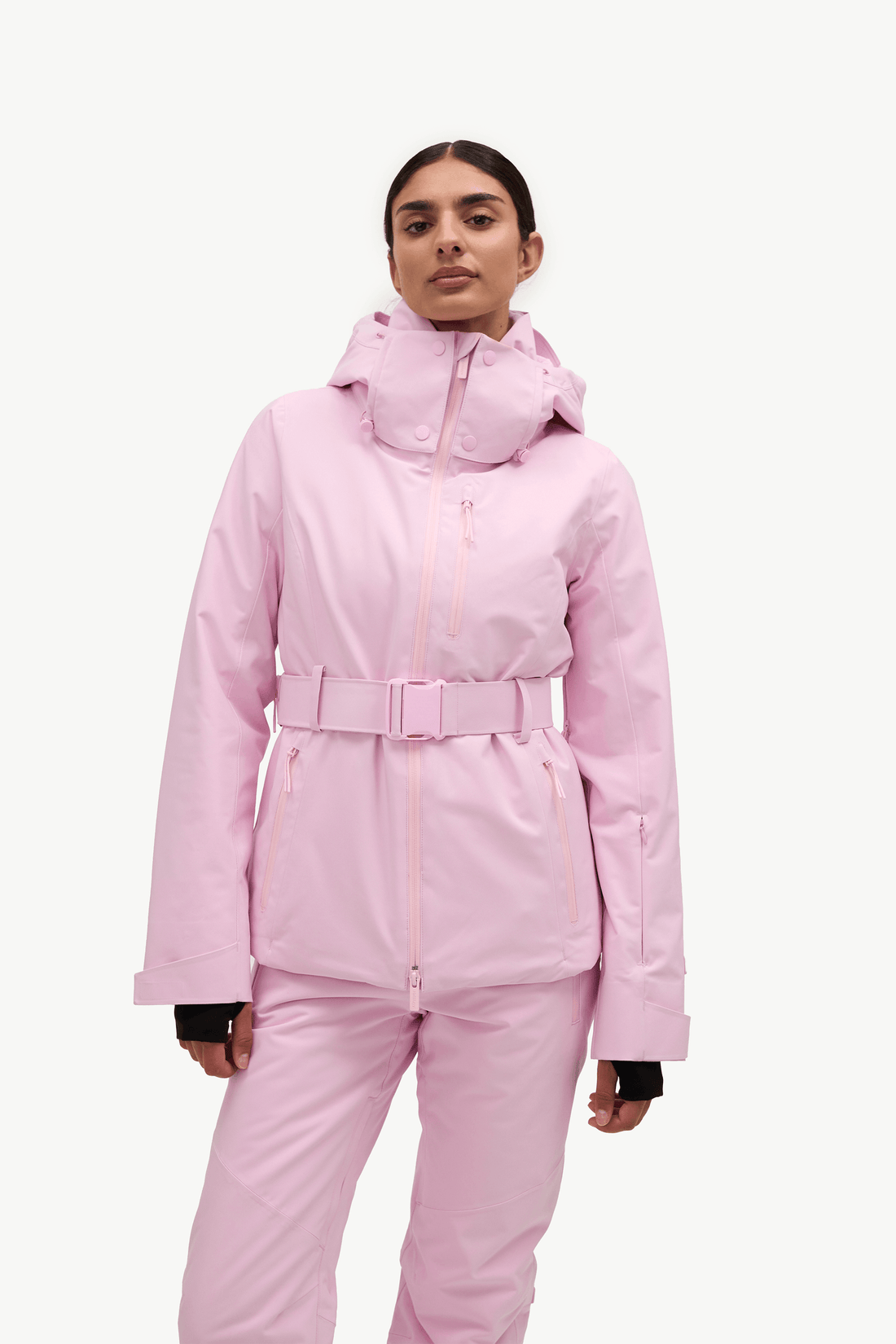 A woman in profile wearing a light pink ski jacket and matching ski pants against a plain white studio background.