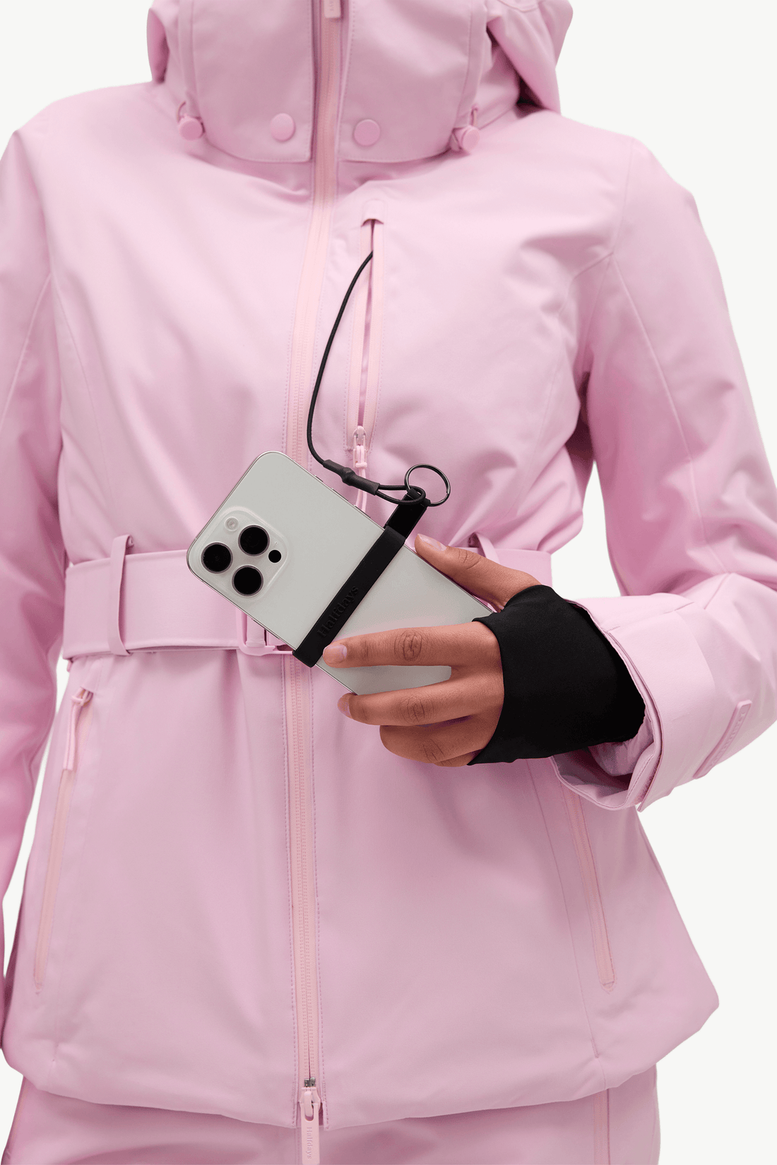 A close-up of a person wearing a light pink hooded ski jacket and matching pants, holding a white smartphone with a black wrist strap attached near the chest.