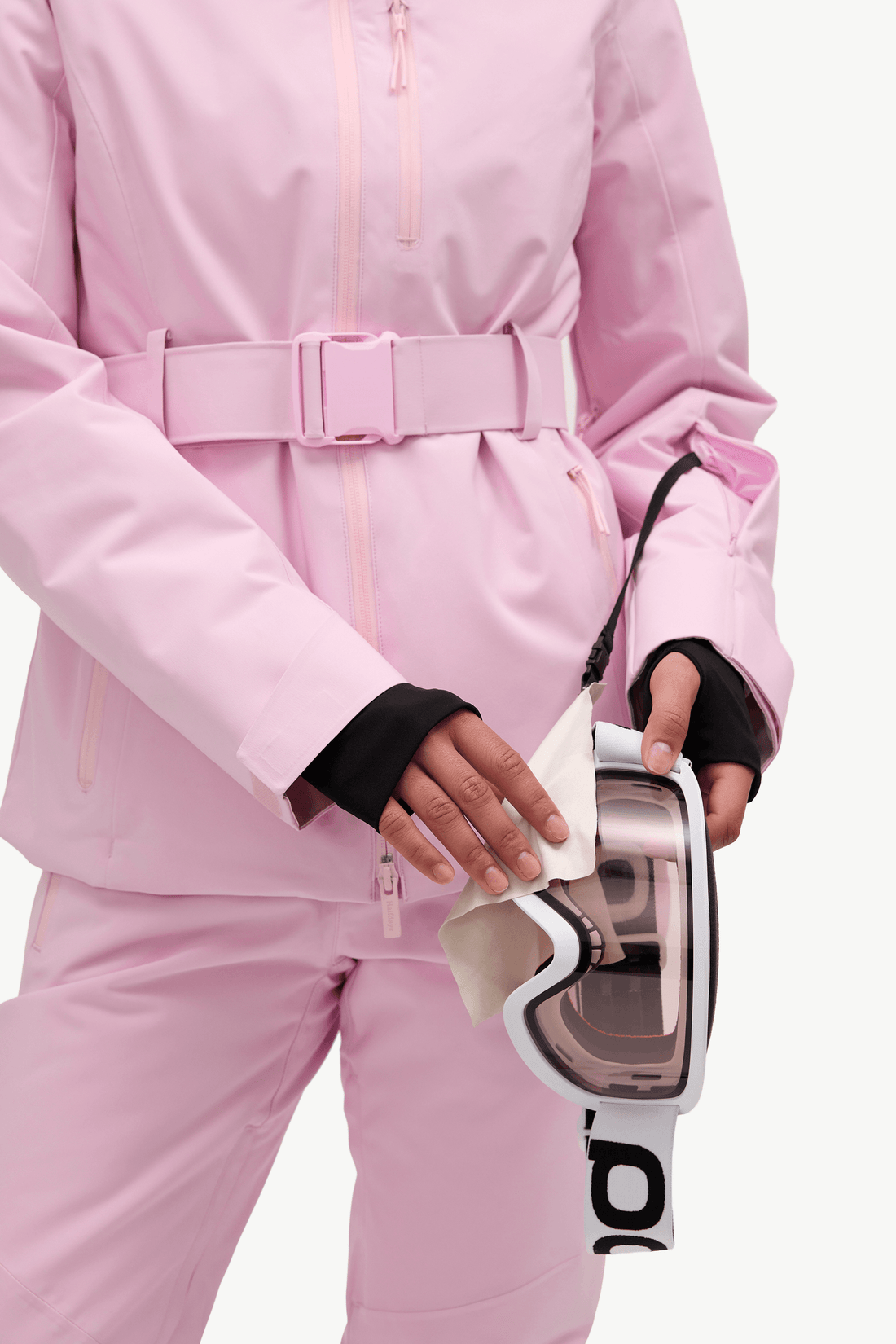A close-up of a person wearing a light pink hooded ski jacket and matching pants, wiping ski goggles with a white cloth.