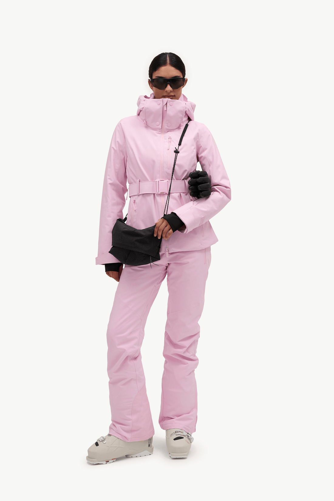 A woman in profile wearing a light pink ski jacket and matching ski pants, with dark gloves and white ski boots, against a plain white studio background.