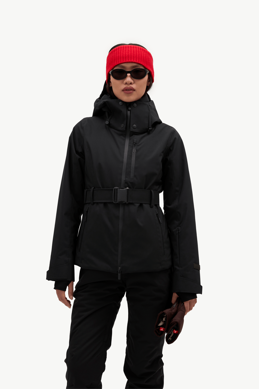 A model wearing a black ski jacket with a matching black belt, black pants, a bright red knit beanie, and black sunglasses, standing against a solid white background.